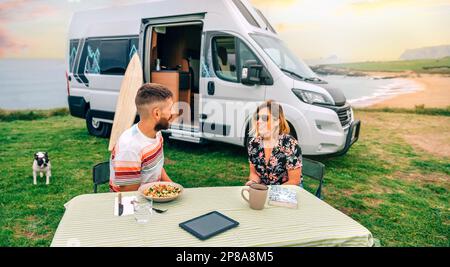 Coppia seduta all'aperto con camper furgone e spiaggia sullo sfondo Foto Stock