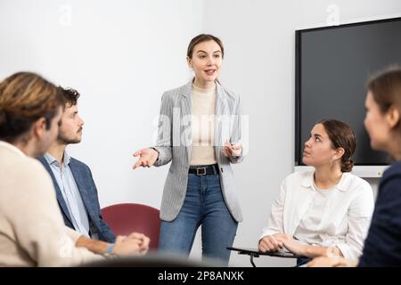 Allenatore professionale donna in piedi vicino a tavola interattiva e condurre formazione diverse persone riunite in cerchio in auditorium Foto Stock