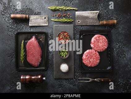 Bistecca di filetto di manzo fresco e hamburger di carne macinata in un vassoio vuoto con sfaldature e rosmarino su sfondo nero con rosmarino. Vista dall'alto. Foto Stock