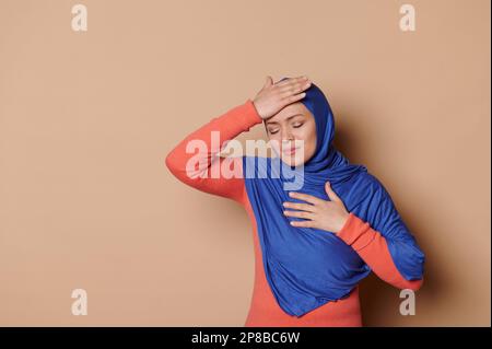 Donna musulmana in hijab blu, tenendo la mano sulla testa, che soffre di mal di testa, terribile emicrania, sfondo beige isolato Foto Stock