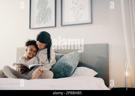 Madre e figlia trascorrono del tempo a leggere un libro insieme, sorridono e si siedono su un letto. La mamma felice sta aiutando il suo bambino con le sue abilità di lettura. Foto Stock