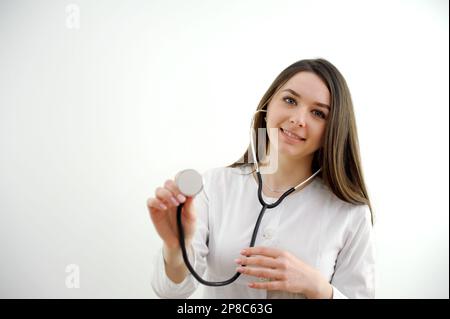 La studentessa medica universitaria impara ad usare correttamente un phonendoscope che tiene nelle sue mani esamina sorridendo in un cappotto bianco sulla formazione di fondo bianco il dottore futuro sia H3althy Foto Stock