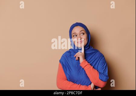 Piacevole donna musulmana positiva in hijab blu, mettendo il dito sul mento, ragionamento, pensiero, sfondo beige isolato Foto Stock