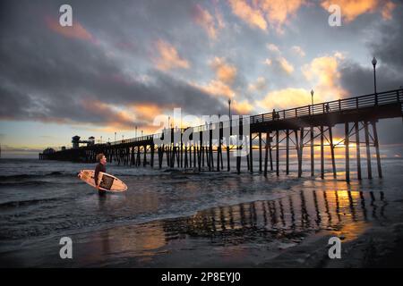 Giovane donna con tavola da surf in piedi presso il molo al tramonto, Oceanside, California, USA Foto Stock