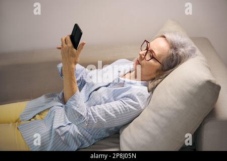 Donna anziana che riposa a casa guarda qualcosa sul suo telefono Foto Stock