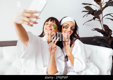 Madre e figlia teen hanno togeather giorno di bellezza, facendo selfi, copia spazio. Madre e figlia usano collagene cerotti d'oro e verde occhio, skincare b Foto Stock