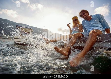 giovane coppia caucasica adulta seduta sul molo che spruzzi d'acqua con piedi nudi Foto Stock