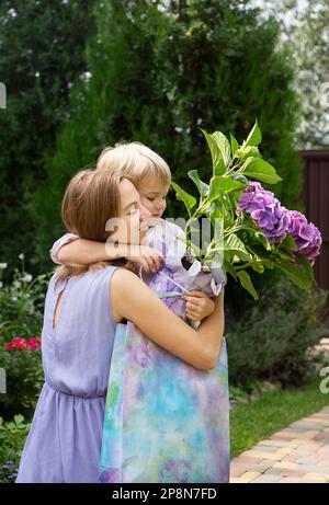 Buona festa della mamma. piccolo figlio si congratula con sua madre in vacanza dà fiori di ortensia viola, regalo e abbracci dolcemente. umore festoso, gratitudine. BIRT Foto Stock