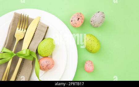 Impostazione tavolo di Pasqua, decorazione uova di Pasqua, tovagliolo e posate dorate su piatti bianchi, sfondo verde pastello Foto Stock