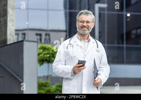 Ritratto di un medico di successo, un uomo più vecchio dai capelli grigi in un cappotto bianco che si trova fuori della clinica e che utilizza il telefono. Guarda la telecamera sorridendo. Foto Stock