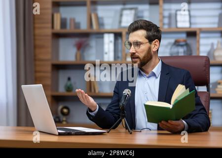 Presentazione online del libro. Un giovane scrittore, autore in tuta, siede in ufficio a un tavolo di fronte a una fotocamera, un computer portatile e un microfono, tiene un libro, legge agli ascoltatori durante una videochiamata. Foto Stock