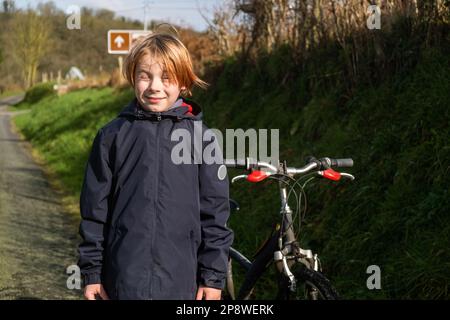 Giovane ragazzo in bicicletta in campagna. Ragazzo di nove o dieci anni di età si trova su erba verde vicino alla strada. Nelle vicinanze c'è la bicicletta. Ricreazione attiva, c Foto Stock