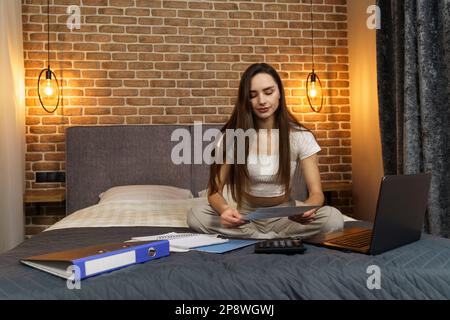 Una giovane donna siede su un letto, guarda i documenti di segnalazione, conta su una calcolatrice e lavora su un computer portatile. Foto Stock