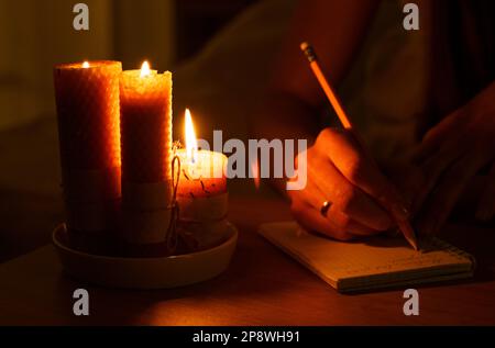 Una donna prende appunti in un notebook in una stanza buia con candele accese. Primo piano. Magia e occultismo. Foto Stock