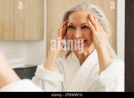 Donna anziana felice con i capelli grigi che guardano uno specchio e che toccano il suo volto mentre guarda lo specchio Foto Stock