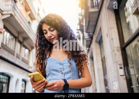 ritratto ad angolo basso di un'attraente donna dai capelli scuri che cammina per la città mentre usa il suo telefono Foto Stock