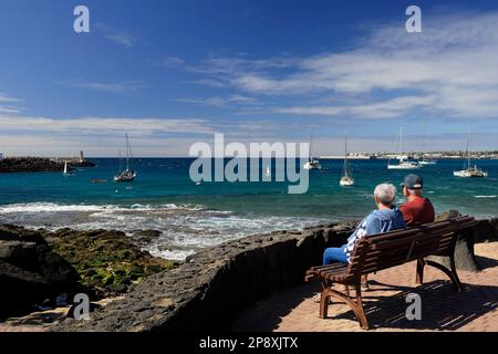 Coppia matura seduta su una panchina che guarda al mare con barche e cielo blu. Lanzarote. Data febbraio / marzo 2023. Foto Stock