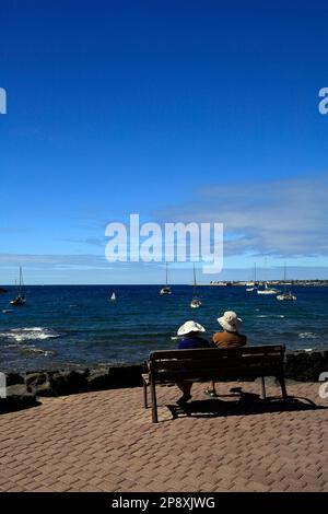 Coppia matura seduta su una panchina che guarda al mare con barche e cielo blu. Lanzarote. Data febbraio / marzo 2023. Foto Stock