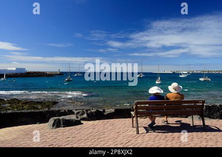 Coppia matura seduta su una panchina che guarda al mare con barche e cielo blu. Lanzarote. Data febbraio / marzo 2023. Foto Stock