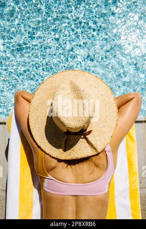 Una bella donna in vacanza si posa su una chaise longue accanto ad una piscina blu con un cappello da sole. Sta riposando la testa nelle sue braccia incrociate guardando fuori. Foto Stock