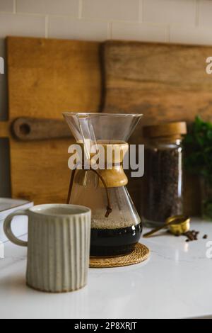 Postazione caffè con chicchi freschi e Chemex con tazza in ceramica sul bancone della cucina Foto Stock