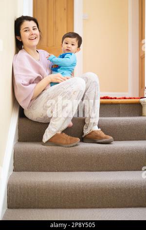Una nuova madre per la prima volta che porta il suo bambino maschile multirazziale seduto in cima alla scala a casa. Guardando e macchina fotografica e ridendo con un grande s Foto Stock