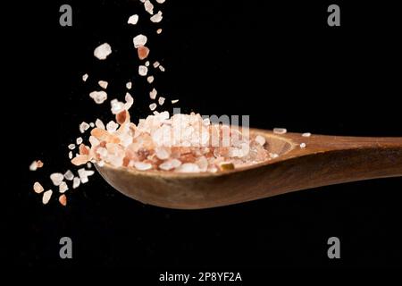 Una foto ravvicinata un sale marino himalayano rosa grossolano che cade sul cucchiaio di legno posto su uno sfondo nero. Foto Stock