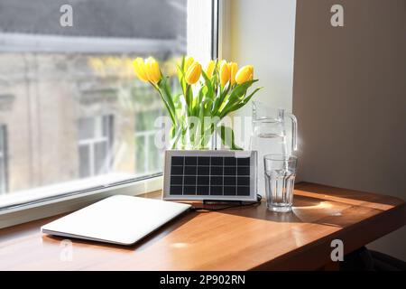Portatile pannello solare di carica computer portatile, caraffa d'acqua e vaso con tulipani sul tavolo vicino alla finestra Foto Stock