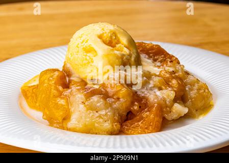 Vista del succoso e delizioso ciottolo di pesche fatto in casa con una paletta di gelato alla vaniglia in un piatto di ceramica bianca su un asse di legno. Foto Stock