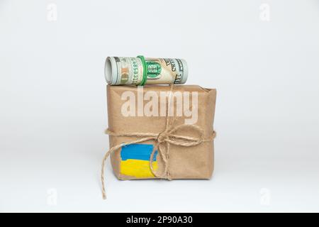Scatola marrone con una bandiera dipinta dell'Ucraina e cento dollari su sfondo bianco, aiuto umanitario per l'Ucraina 2023 Foto Stock