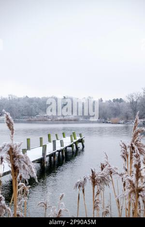 Pontile in legno coperto di neve in lago d'acqua ghiacciato e calmo con canne d'arancio in primo piano Foto Stock