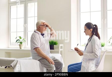 Un paziente anziano che si lamenta di mal di testa a un medico donna attento in un moderno ospedale clinico. Foto Stock