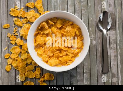 Vista dall'alto di appetitosi corn flakes in una ciotola bianca con latte posto su un tavolo di legno a strisce vicino al cucchiaio Foto Stock