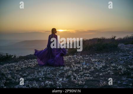 Tramonto viola abito donna montagne. Ascesa del mistico. tramonto sulle nuvole con una ragazza in un lungo vestito viola. Nel prato c'è un prato Foto Stock