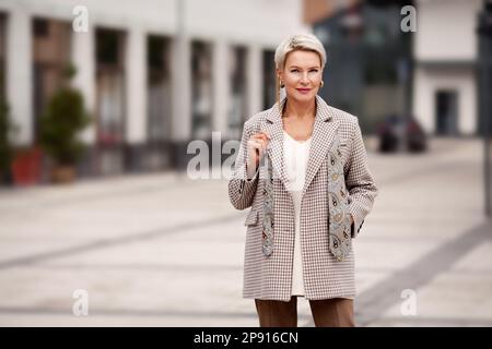 Ritratto di bella fidata donna matura business con bionda taglio corto fashionably vestito in giacca doppia breasted e cravatta, in piedi sul str Foto Stock