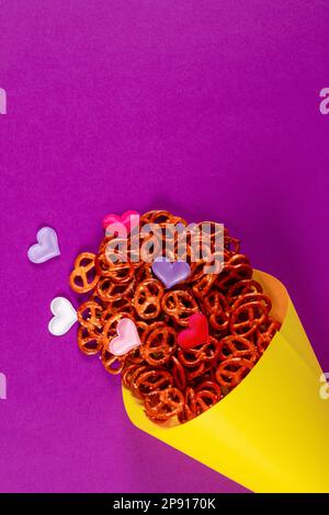 Piccoli pretzel e piccoli cuori colorati in cono di carta giallo giallano su sfondo viola con spazio negativo per il posizionamento della copia. Pretzel domenica concetto ba Foto Stock