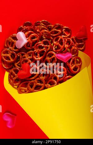 Piccoli pretzel e piccoli cuori colorati in cono di carta giallo si trovano su sfondo rosso primo piano. Pretzel domenica concetto Foto Stock