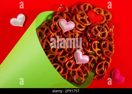 Mini pretzel e piccoli cuori colorati in cono di carta verde chiaro giacciono su sfondo rosso primo piano. Pretzel domenica concetto Foto Stock