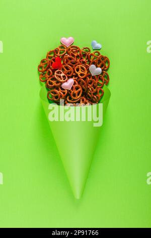 Piccoli pretzel e piccoli cuori colorati in cono di carta verde chiaro giacciono su sfondo verde brillante. Luminoso concetto di Oktoberfest Foto Stock