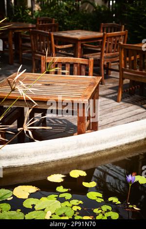 Patio con caffè all'aperto con vecchi tavoli e sedie in legno scabrido alla luce del sole. Laghetto con gigli d'acqua su una romantica terrazza ristorante Foto Stock