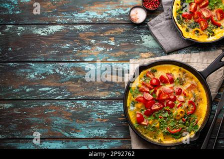 Frittata sana in due padelle di ghisa con uova sbattute fritte e verdure di stagione su sfondo rustico in legno. Omelette italiana con biologico Foto Stock