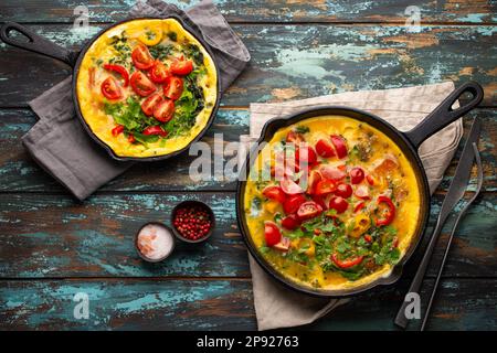 Frittata sana in due padelle di ghisa con uova sbattute fritte e verdure di stagione su sfondo rustico in legno. Omelette italiana con biologico Foto Stock