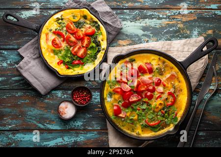 Frittata sana in due padelle di ghisa con uova sbattute fritte e verdure di stagione su sfondo rustico in legno. Omelette italiana con biologico Foto Stock