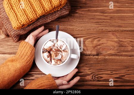 Accogliente piatto invernale, mano femmina che tiene tazza con cioccolata calda o cacao con marshmallow e pile di caldi vestiti a maglia su rustico di legno Foto Stock