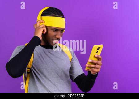 Uomo etnico nero con zaino e cuffie gialle su sfondo viola, concetto studente, con il telefono serio Foto Stock