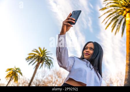 Giovane donna africana che posa in abiti bianchi in un luogo tropicale con palme al tramonto godendosi l'estate prendendo un selfie Foto Stock