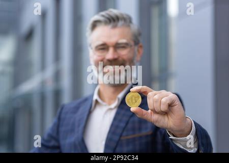Primo piano. Ritratto di felice uomo anziano dai capelli grigi, uomo d'affari in piedi fuori ufficio e tenendo in mano la moneta d'oro. Presenta e mostra il bitcoin alla fotocamera. Foto Stock