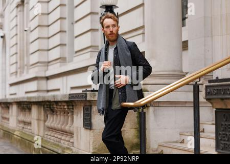 Uomo elegante barbuto in una giacca e sciarpa scende le scale lasciando l'edificio Foto Stock