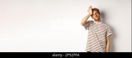 Giovane uomo stanco con capelli rossi e occhiali, occhi rotolanti e facepalm fastidio, in piedi su sfondo bianco Foto Stock