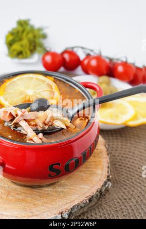 Solyanka di carne. Caldo combinato hodgepodge. una porzione di zuppa di carne rossa piccante con pomodori, salsicce affumicate e olive al limone in una turena rossa Foto Stock